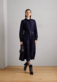 Navy blauwe lange mouwen jurk met knoopsluiting aan de voorkant en een ceintuur in de taille. Glanzende zwarte enkellaarsjes en een kleine zwarte handtas maken de outfit compleet.