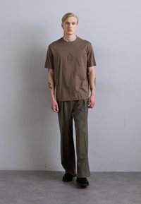 Camiseta marrón de manga corta con un sutil detalle de logo, combinada con pantalones de pierna ancha en color verde oliva y zapatos negros, todo sobre un fondo neutro.