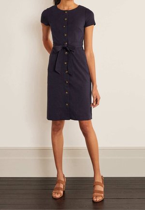 Vestido azul marino con mangas cortas, escote redondo y botones en la parte delantera. Presenta un detalle de cinturón anudado y un dobladillo a la altura de la rodilla. Se muestran sandalias marrones de tacón.