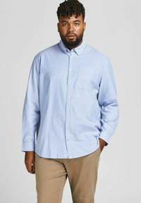Camisa de botón de manga larga de color azul claro, hecha de una tela texturizada, que presenta un bolsillo en el pecho y un collar, combinada con pantalones caqui.