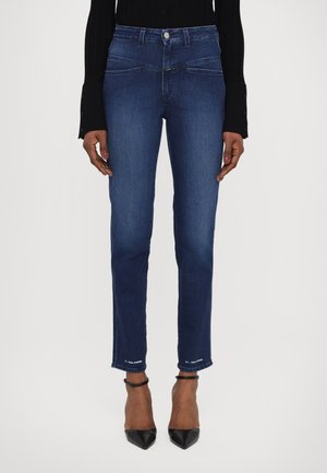 Nogi w ciemnoniebieskich obcisłych dżinsach z nogawkami sięgającymi kostek, zestawione z czarnymi szpilkami z ostrym noskiem i czarnym topem z długim rękawem.