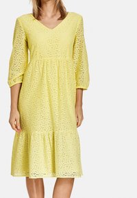 Vestido de encaje amarillo con escote en V, mangas abullonadas de tres cuartos y dobladillo acampanado. Presenta intrincados patrones florales por todo el tejido.