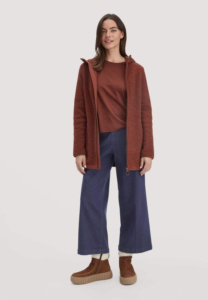 Roodbruine fleece jack met capuchon, gecombineerd met een bijpassend topje en brede spijkerbroek. Bruine enkellaarsjes met gestructureerde zolen.