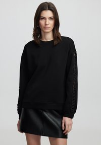 Un sweat-shirt noir avec col rond, coupe ample et détails tressés le long des manches, associé à une mini-jupe en cuir noir.