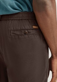 Braune leichte Hose mit elastischem Bund, ausgestattet mit einer hinteren Tasche mit Knopfverschluss und einem kleinen Etikett.