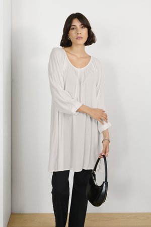 Tunique crème légère à manches longues avec encolure dégagée et détails froncés. Assortie à un pantalon noir ajusté et à un sac à main noir.