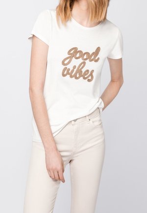 Kobieta w białej koszulce z krótkim rękawem z napisem „good vibes” w beżowej kursywie oraz jasnobeżowych spodniach, pozująca z jedną ręką w kieszeni.