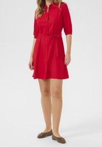 Czerwona sukienka koszulowa z krótkim rękawem, zapinana na guziki, z paskiem w talii. Wykonana z gładkiego materiału, posiada rozkloszowaną spódnicę oraz delikatnie wykończone mankiety.
