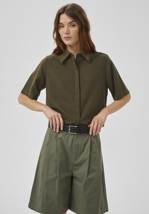 Vrouw die een zwarte riem verstelt, gekleed in een olijfgroene shirt met korte mouwen en hooggetailleerde, wijdvallende shorts tegen een effen achtergrond.