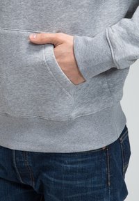 Hand in der vorderen Tasche eines grauen Sweatshirts, das mit dunkelblauen Jeans getragen wird.