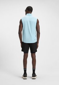 Čovjek odjeven u svijetloplavu košulju bez rukava, crne kratke hlače, crne čarape s natpisom "BOSS" i crne tenisice, stoji okrenut leđima na jednostavnoj bijeloj pozadini.