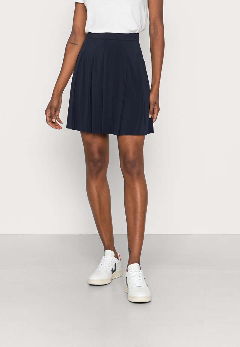 Femme portant un haut blanc, une mini-jupe plissée bleu marine et des baskets blanches, debout devant un fond clair uni.