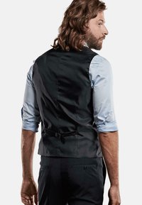 Schwarze Weste mit glatter Textur und verstellbarem Riemen am Rücken, getragen über einem hellblauen Hemd mit hochgekrempelten Ärmeln. Maßgeschneiderte Passform, elegantes Design.