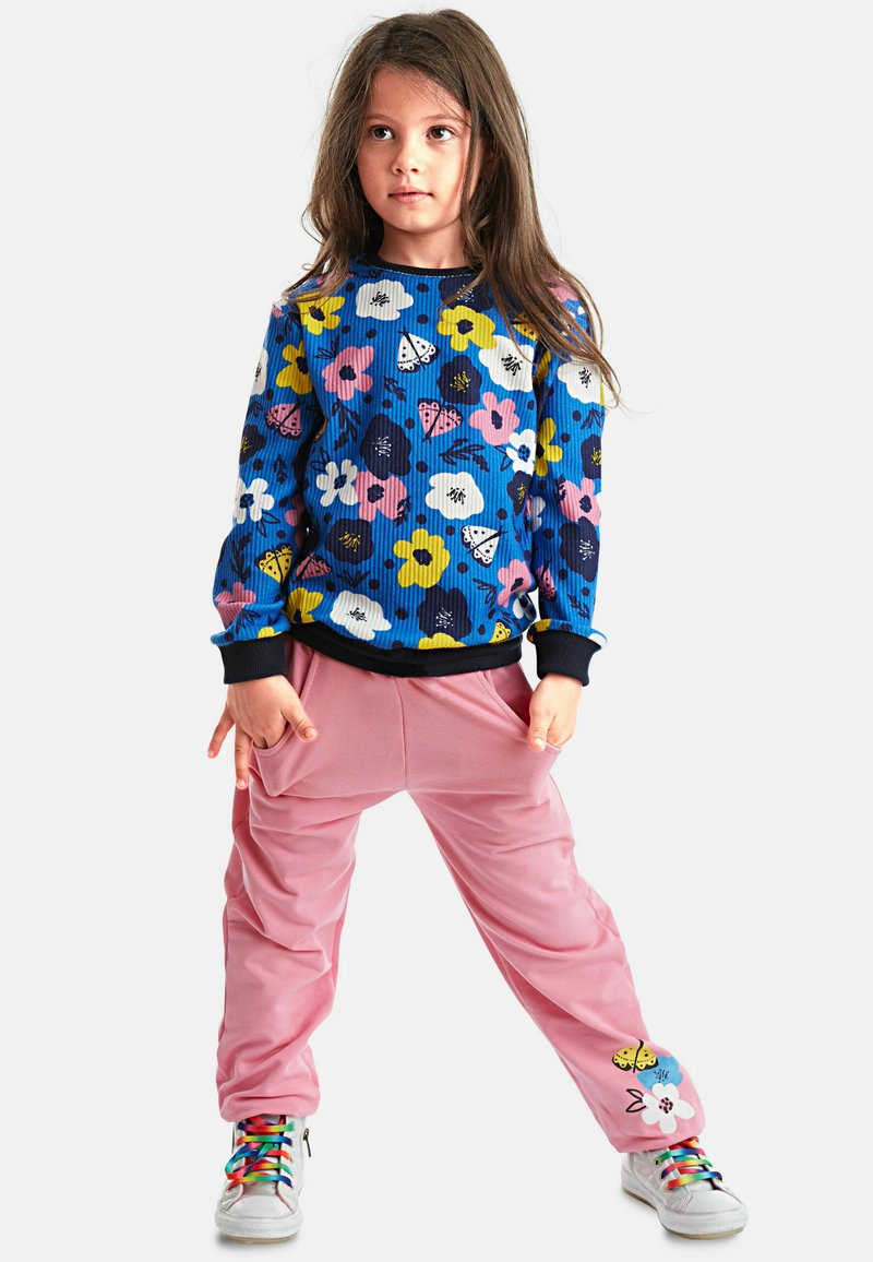 Langärmliger blauer Rippstrickpullover mit mehrfarbigem Blumendesign, kombiniert mit locker sitzenden rosa Hosen mit Blumenakzenten und bunten Schuhen.