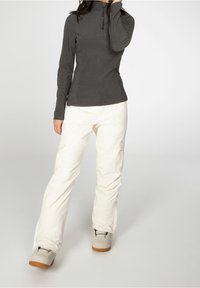 Kvinde iført en mørkegrå langærmet bluse, hvide cargo-bukser og beige sneakers, der står mod en ensfarvet lys baggrund.