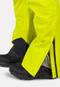 Pantaloni impermeabili gialli con accenti neri, dotati di aperture con zip alle caviglie, abbinati a scarpe nere robuste.