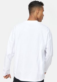 Camisa blanca de manga larga hecha de algodón. Presenta un escote redondo y un ajuste relajado. La vista trasera muestra un sutil logotipo cerca del cuello.