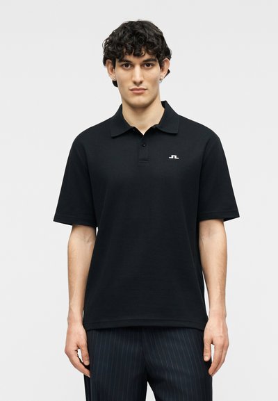 Jeune homme portant un polo à manches courtes noir avec un petit logo blanc sur la poitrine et un pantalon foncé à fines rayures.