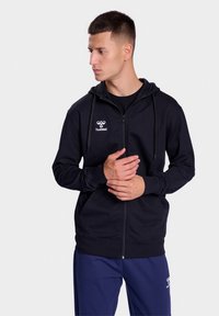 Zwarte rits hoodie van zacht materiaal, met een logo op de borst en een capuchon met koord, voorzien van zijzakken en ribgebreide manchetten.