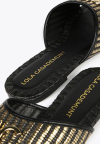 Black and gold woven slide sandals with a flat sole, featuring a braided edge and a logo in gold on a black footbed.