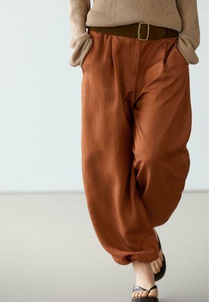 Persona che indossa pantaloni larghi color ruggine con una cintura marrone, maglione beige e sandali neri con punta aperta, con le mani nelle tasche, camminando in avanti.
