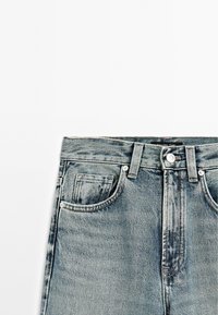 Blue denim jeans waist section showing button, belt loops, front pockets, and faded fabric texture on white background.