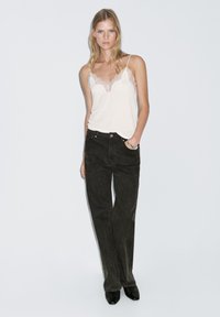 Weißes Trägeroberteil mit Spitzenbesatz, kombiniert mit hochgeschnittenen, dunkelgrünen Schlagjeans. Das Outfit wird von eleganten schwarzen Absätzen ergänzt.