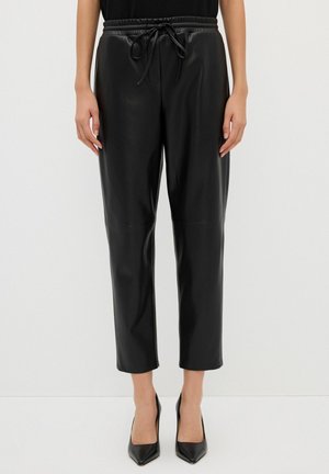 Kobieta ubrana w czarne proste spodnie z ekoskóry z elastycznym ściągaczem w talii i czarne buty na obcasie z ostrym noskiem, stoi na białym tle.