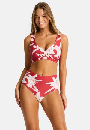 Uśmiechnięta kobieta w czerwonym kwiecistym bikini z wysokim stanem i skręconym przodem, stojąca na jasnym, jednolitym tle.