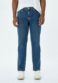 Jeans de mezclilla azul con un corte recto, cinco bolsillos, cierre de botón de metal y un ligero desgaste en la tela. Combinados con zapatillas blancas.