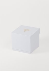 Hvid tekstureret terningformet kasse med et guld Guess trekantet logo på toppen, placeret mod en ensartet hvid baggrund.