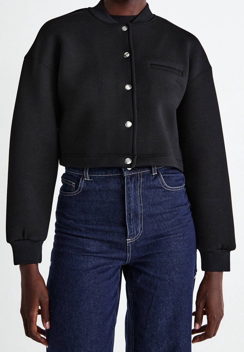 Personne portant une veste noire courte boutonnée avec des manches longues et un jean bleu foncé taille haute sur un fond uni.