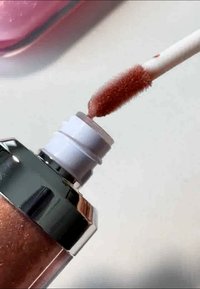 Tubo de brillo labial con base metálica plateada, parte superior cilíndrica transparente y un aplicador de textura suave, recubierto en un color rosa brillante.