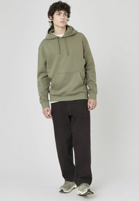 Haut à capuche vert olive avec une poche avant et un cordon de serrage, associé à un pantalon noir ample et des chaussures de sport grises.