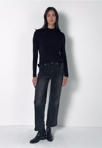 Woman standing against white wall wearing black long-sleeve sweater, black jeans, and black ankle boots, facing forward.