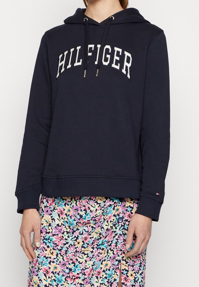 Personne portant un sweat à capuche bleu marine avec le texte "HILFIGER" et une jupe à motifs floraux avec des fleurs colorées et une fente à l'avant.