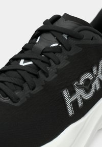 Μαύρο αθλητικό παπούτσι με μαύρα κορδόνια, υφή στην επάνω επιφάνεια και το λογότυπο «HOKA» στο πλάι, με χοντρή λευκή σόλα.