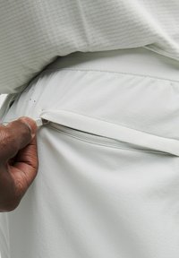 Weiße Sportshorts aus strukturiertem Stoff mit einem elastischen Bund und einer seitlichen Reißverschlusstasche, dargestellt mit einer Hand, die den Reißverschluss greift.