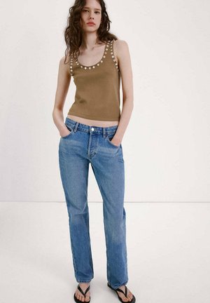 Jeune femme portant un haut marron sans manches avec des détails œillets, un jean bleu taille haute et des sandales noires, debout les mains dans les poches.