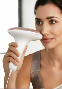 Appareil d'épilation Philips, avec un corps blanc, des accents roses, une forme ergonomique et un design avec cordon, tenu près du visage de l'utilisateur.