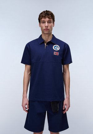 Mand iført navy polo shirt med mærker og matchende navy shorts mod en ensartet lys baggrund, stående med armene langs siden.