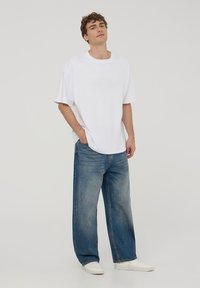 T-shirt bianca oversize con maniche corte, abbinata a jeans baggy blu chiaro e sneakers bianche. Tessuto morbido e casual con un fit rilassato.