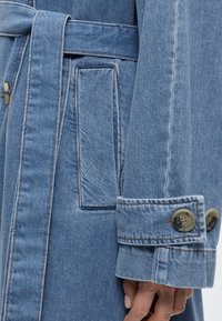 Artimas mėlyno džinsinio paltuko rankovės vaizdas su sagomis užsegamu rankogaliu, diržo kilpa ir šonine kišene, dėvimas asmens su matomais pirštais.