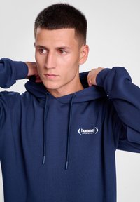 Marinblå hoodie i mjuk tyg, med en känguruficka, dragsko för huvan och en vit logotyp på vänster bröstområde.