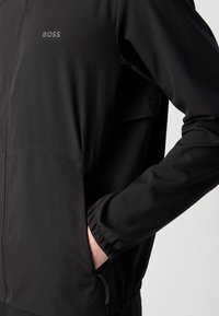 Čierna bunda na zips s elastickými manžetami a bočnou zipsovou vreckom, ruka vložená do vrecka, logo "BOSS" na ľavom hrudníku.