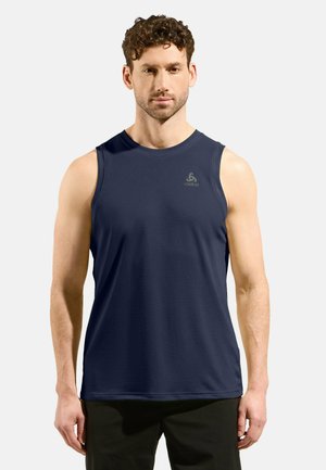 Mann mit lockigem Haar, der ein dunkelblaues ärmelloses Sportshirt und schwarze Shorts trägt, steht vor einem einfarbigen hellen Hintergrund.