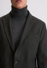 Donkergrijze blazer met een subtiele textuur, enkele knoopsluiting en notch revers, gedragen over een donkere coltrui.