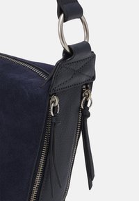 Sac à bandoulière en cuir suédé bleu marine et en cuir grainé avec des accessoires en métal argenté, fermeture éclair, et des détails de pompons sur les tirettes de fermeture.