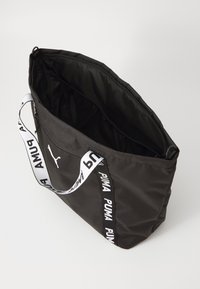 Svart tote-väska med dragkedjestängning och en framträdande vit PUMA-logotyp längs axelremmen, sedd ovanifrån.