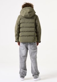 Chaqueta de plumas verde oliva con capucha, que presenta un acolchado en forma de rombos y puños elásticos, combinada con jeans sueltos de color gris claro y zapatillas blancas.